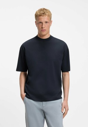Jeune homme aux cheveux blonds courts, portant une chemise noire à manches courtes et un pantalon gris clair, debout les mains dans les poches sur fond blanc.