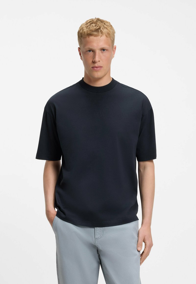 Jeune homme aux cheveux blonds courts, portant une chemise noire à manches courtes et un pantalon gris clair, debout les mains dans les poches sur fond blanc.