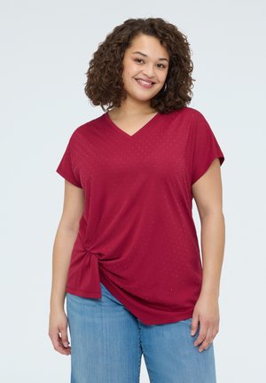 Lächelnde Frau mit lockigem Haar, die eine rubinrote, kurzärmelige V-Ausschnitt-Bluse mit seitlichem Knotendetail und hellblaue Jeans trägt.