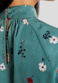 Blusa verde acqua con stampa floreale, dotata di chiusura posteriore con bottoni, dettagli arricciati e tessuto morbido e testurizzato.