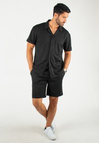 Chemise noire à manches courtes avec des boutons, assortie à un short noir. L'ensemble présente une coupe décontractée et une texture de tissu lisse, complété par des baskets blanches.