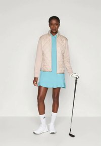 Ženska v svetloplavih golf oblačilih in bež prešit vest stane ter drži golf palico, oblečena v bele golf rokavice, nogavice in teniske.