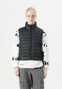 Crna umjetna prsluk s visokim ovratnikom, složen preko bijele flis veste s uzorkom crnog teksta. Sive cargo hlače upotpunjuju outfit.