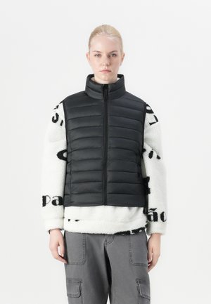 Chaleco acolchado negro con cuello alto, sobre un suéter de forro polar blanco que presenta patrones de texto en negro. Pantalones cargo grises completan el atuendo.