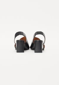 Une paire de sandales à talons ouverts noires avec de larges talons blocs et des sangles élastiques, montrées de l'arrière sur fond blanc.