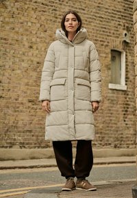 Beige polstret lang frakke med hætte, der har frontknapper, to store lommer og et quiltet mønster, parret med brune sweatpants og sko.