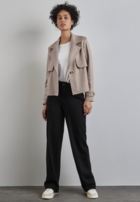 Beżowa krótka kurtka z dużymi przednimi kieszeniami i zapięciem na jeden guzik, zestawiona z czarnymi spodniami z wysokim stanem i białymi sneakersami.