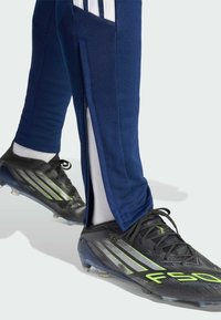 Pantalones deportivos azul marino con franjas blancas laterales, bolsillos laterales con cremallera y ajuste cónico, combinados con botas de fútbol negras con detalles verdes.