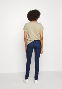 Vaaleanbeige lyhythihainen t-paita yhdistettynä tummansinihin slim fit -farkkuihin ja valkoisiin lenkkareihin, nähtynä takaa valkoista taustaa vasten.