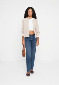 Light beige cardigan with round neckline and buttons, paired with a white crop top and blue straight-leg jeans. Brown handbag and dark shoes.