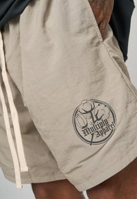 Bermudas beige hechas de tela ligera, con cordón y un logotipo gráfico negro con elementos florales en la pierna izquierda.