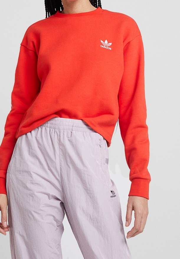 Červená mikina s dlouhým rukávem a kulatým výstřihem, na které je bílé logo Adidas na hrudi, v kombinaci s lehkými kalhotami světle fialové barvy s logem Adidas.