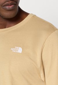 Μπεζ μακρυμάνικο πουκ άμισο με στρογγυλή λαιμόκοψη και μικρό λευκό κεντητό λογότυπο στο αριστερό στήθος, κατασκευασμένο από απαλό βαμβακερό ύφασμα.