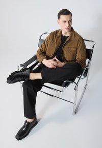 Braune Cordjacke mit zwei Brusttaschen, schwarzes T-Shirt, schwarze Hose und glänzende schwarze Schuhe, sitzend auf einem Chromstuhl vor einem hellen Hintergrund.