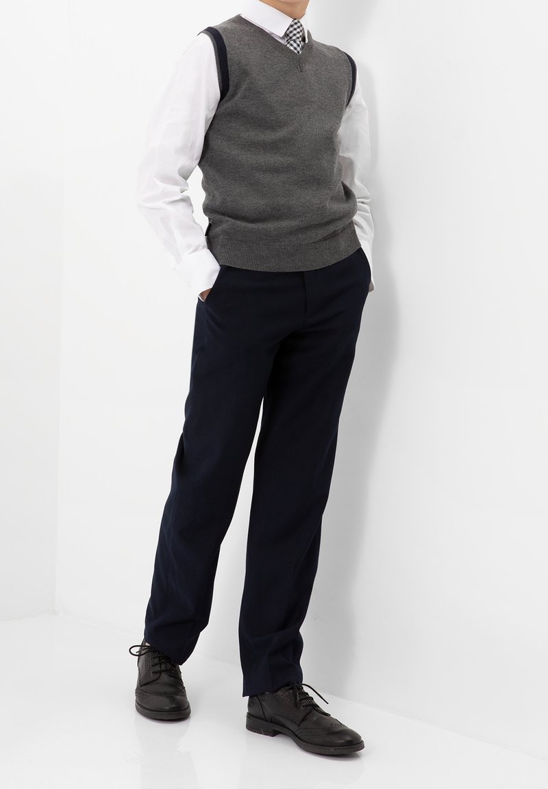 Gilet en laine gris sur une chemise blanche, associé à un pantalon bleu marine foncé et des chaussures en cuir noir. Le gilet présente un col en V et des bords côtelés.