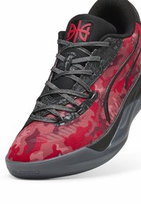 Baskets de sport rouges et noires avec un motif camouflage, tissu texturé, forme peu conventionnelle, et des détails noirs élégants sur la semelle et le matériel.