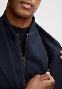 Granatowa kurtka na zamek błyskawiczny o gładkiej fakturze, z prążkowanym kołnierzem i kontrastową podszewką. Posiada widoczny zamek z eleganckim suwakiem.