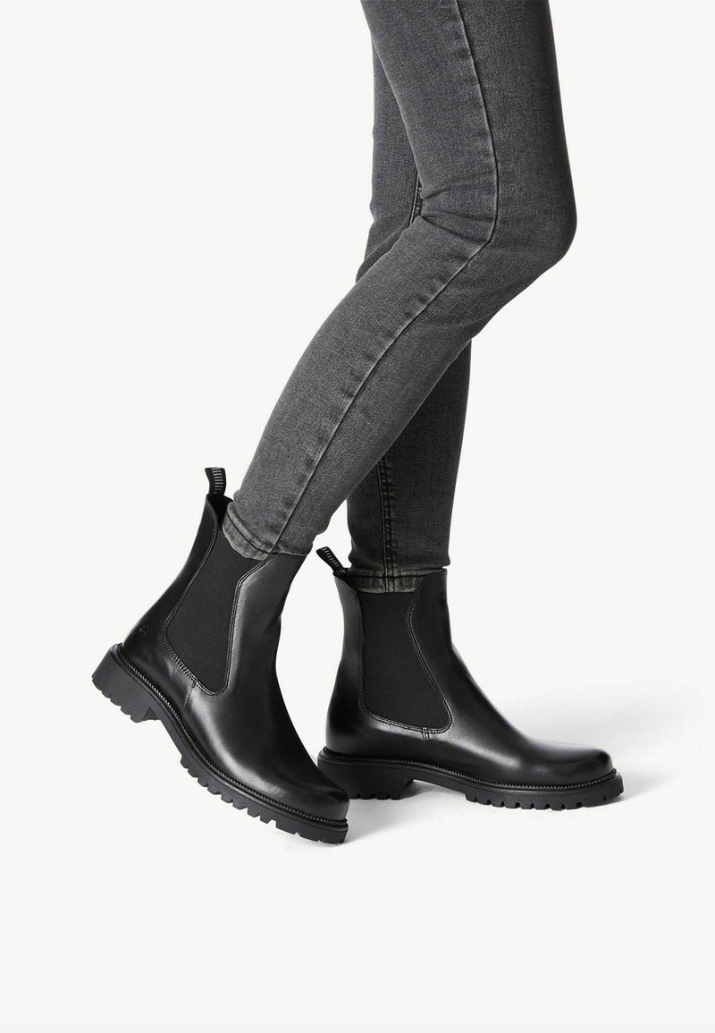 Bottes Chelsea noires avec des panneaux latéraux élastiques, bout rond et semelle en caoutchouc texturée, associées à un jean gris ajusté.