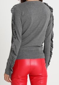 Pull gris avec des accents froncés le long des manches, texture côtelée et coupe ajustée. Associé à un pantalon rouge brillant pour créer un contraste.