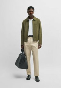 Olivengrön mockajacka över en vit t-shirt, beige byxor och svarta läderskor, håller en stor svart duffelväska.