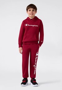 Felpa con cappuccio rossa e pantaloni della tuta abbinati con il logo "Champion" bianco. Il tessuto sembra morbido. Sneakers nere e rosse completano l'outfit.
