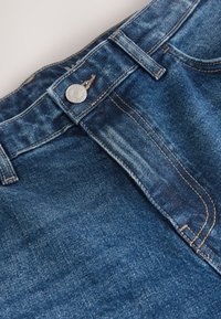 Gros plan sur un jean en denim bleu montrant la ceinture, le bouton en métal, les passants de ceinture et les coutures de la poche avant.