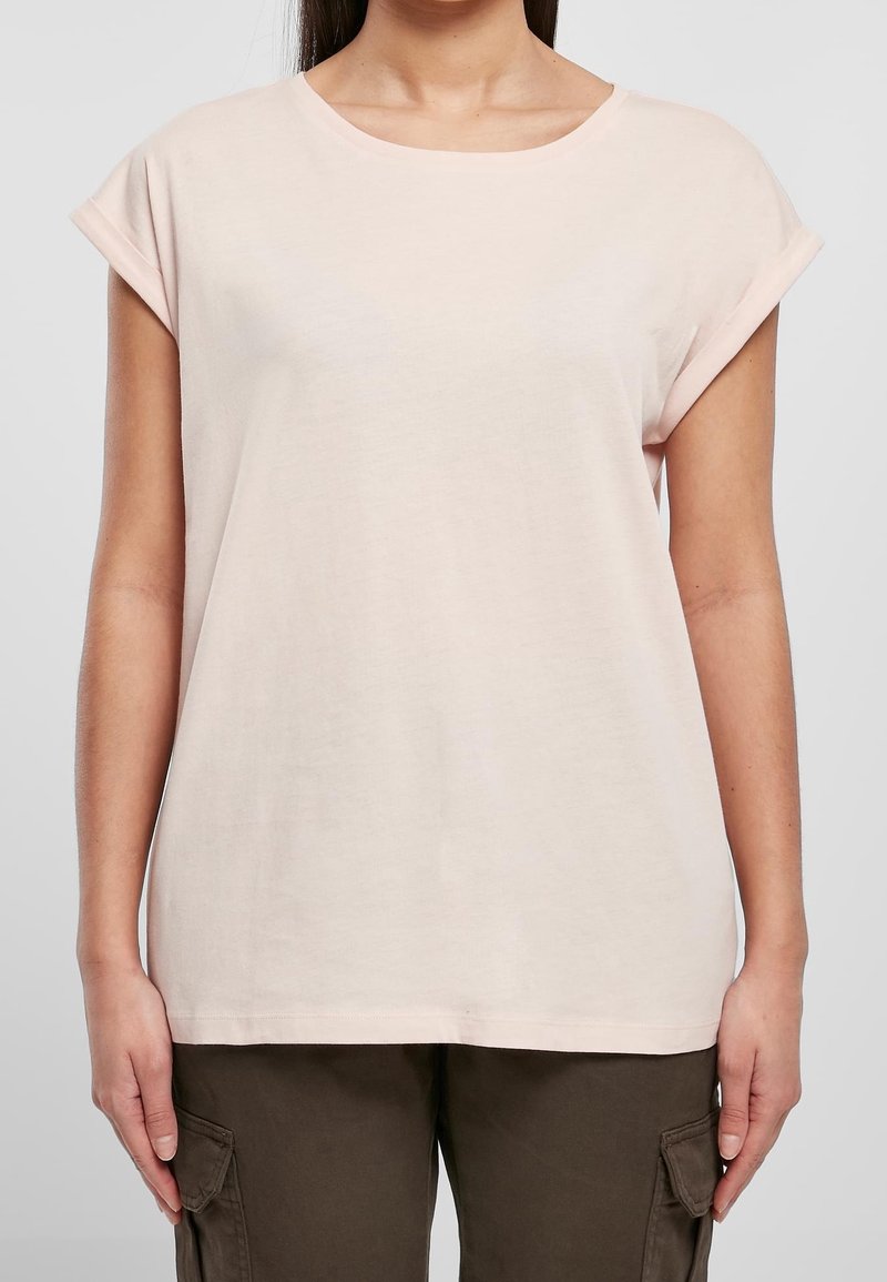 Osoba w jasnoróżowej koszulce z krótkim rękawem i podwiniętymi mankietami oraz ciemnooliwkowych spodniach cargo na jednolitym tle.