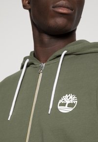 Sudadera con cremallera en verde oliva, con cordones blancos y logo. Presenta una textura suave y una cremallera frontal, mostrando un gráfico sencillo de un árbol.