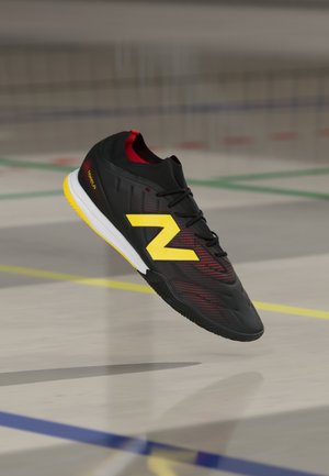 Zapato de fútbol sala negro y rojo con un gran logo amarillo en forma de "Z" flotando sobre un suelo de gimnasio marcado con líneas de colores.