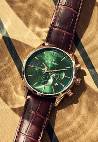 Montre chronographe avec un cadran vert, des accents dorés et des chiffres rouges. Elle dispose d'un bracelet en cuir avec un motif crocodile brun et d'un affichage de la date.