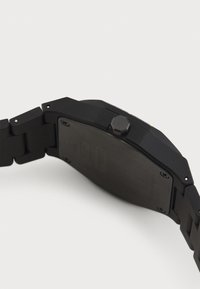 Montre en métal noir avec un bracelet à maillons, couronne hexagonale et un fond de boîtier texturé. Design épuré et angulaire avec peu de détails.