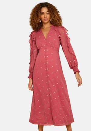 Nő, aki egy hosszú ujjú, poros rózsaszín midi ruhát visel kis szívmintákkal, gombos elülső résszel, fodros vállakkal és ráncolt mandzsettákkal.