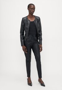 Blouson en simili cuir noir, haut ajusté noir et pantalon noir fuselé. Chaussures pointues avec des pois blancs. Texture lisse dans l'ensemble.