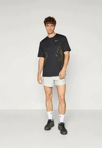 Černé sportovní tričko s žlutými akcenty, krátké rukávy; v kombinaci s světlými šedými šortkami a černými botami. Minimalistický design, tkanina odvádějící vlhkost.