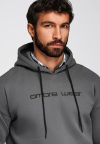 Mężczyzna z ciemnymi kręconymi włosami i brodą nosi szary sweatshirt z napisem "ombre wear", nałożony na koszulę w kratę z kołnierzykiem, na jednolitym tle.