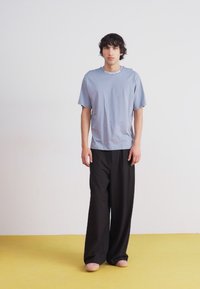 T-shirt azzurro chiaro a maniche corte con scollatura tonda. Abbinato a pantaloni neri larghi e pantofole chiare, su un pavimento giallo.