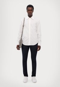 Λευκό πουκάμισο με μακριά μανίκια και κλασικό γι ακά, συνδυασμένο με σκούρους στενούς τζιν και λευκά αθλητικά παπούτσια. Ένα γκρι τσάντα χιαστί κρέμεται από τον ώμο.