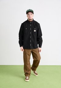Schwarzes Hemd mit Knöpfen und zwei Brusttaschen, braune Cargo-Hosen und braune Sneakers an einer Person vor einem hellen Hintergrund und einem grünen Boden.