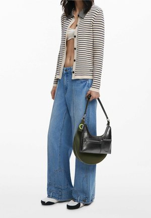 Frau trägt gestreiften Cardigan über weißem Crop-Top, weite blaue Jeans, schwarz-weiße Schuhe, hält schwarze Handtasche mit grünem rundem Boden.