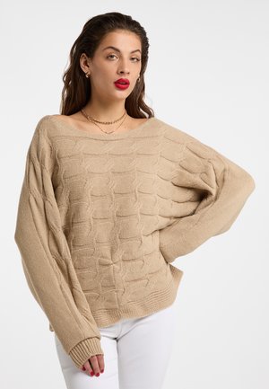 Donna con capelli castani ondulati indossa un maglione beige a trecce e pantaloni bianchi, posando con una mano sui fianchi davanti a uno sfondo bianco.
