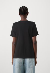 Person med flätat hår som bär en enkel svart t-shirt med kort ärm och ljusblå denimjeans, sedd bakifrån mot en vit bakgrund.