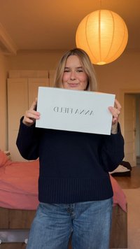 Witte rechthoekige doos met "ANNA FIELD" geschreven in zwarte typografie. De persoon draagt een donkere trui en houdt de doos voor zich.