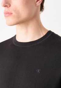 Pull noir à col rond fabriqué en tissu tricot doux, avec un subtil logo "CK" brodé sur la poitrine et des détails côtelés aux poignets et à l'ourlet.