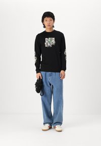 Chemise noire à manches longues avec un design graphique au centre et des accents à motifs sur les manches. Associée à un jean baggy bleu clair et des chaussures.
