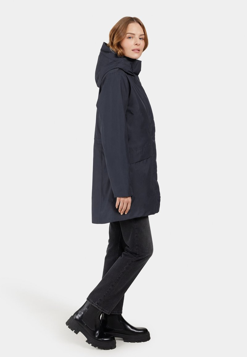Marineblauer wasserdichter Parka mit Kapuze, zwei Vorderaschen, gerader Schnitt und langen Ärmeln, kombiniert mit schwarzen Jeans im geraden Schnitt und klobigen Stiefeln.
