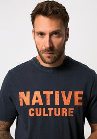 Tmavě námořnické tričko s krátkým rukávem vyrobené z měkké bavlny, s opotřebovaným oranžovým potiskem, který říká "NATIVE CULTURE" napříč hrudníkem.