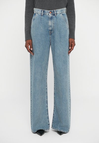 Persoon in lichtblauwe hoge taille, wijde jeans met voorzakken en een oranje knoop, gecombineerd met een donkergrijze lange mouw top.
