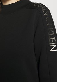 Svart sweatshirt med hög krage och "CALVIN KLEIN" tryckt i silver längs axeln. Fokusera på stil och varumärke.