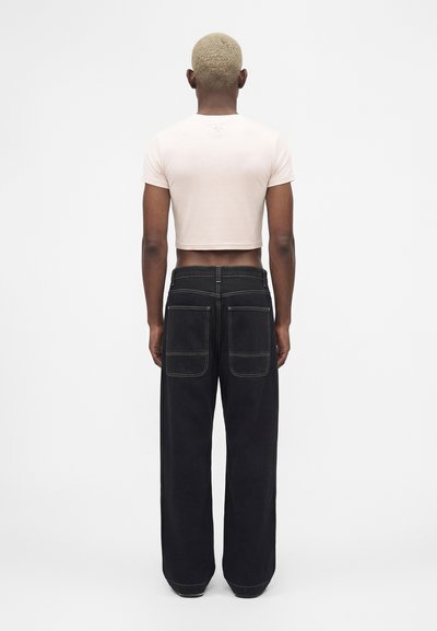Άτομο με κοντά ξανθά μαλλιά που φοράει λευκό κοντό μπλουζάκι και φαρδιά μαύρη παντελόνα με μεγάλες πίσω τσέπες, στέκεται με την πλάτη.