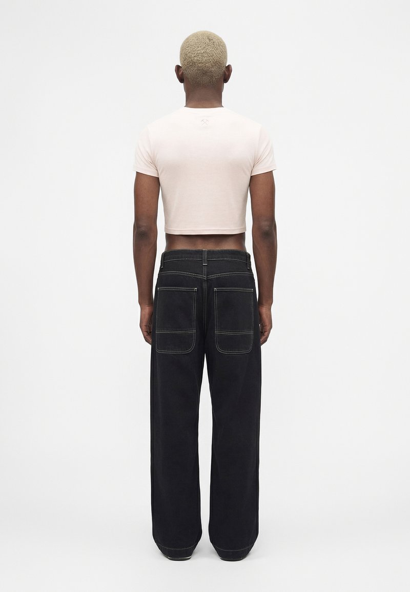 Personne aux cheveux blonds courts portant un t-shirt blanc court et un pantalon noir ample avec de grandes poches arrière, debout de dos.